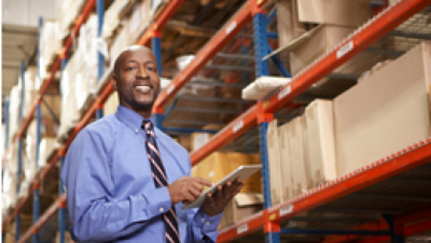 Smiling man in shirt and tie stands next to warehouse shelves.